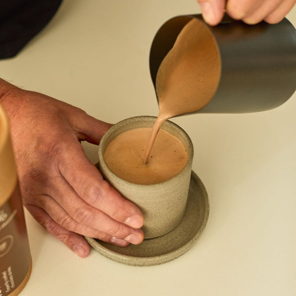 Person pouring a creamy liquid into a ceramic cup on a light surface.