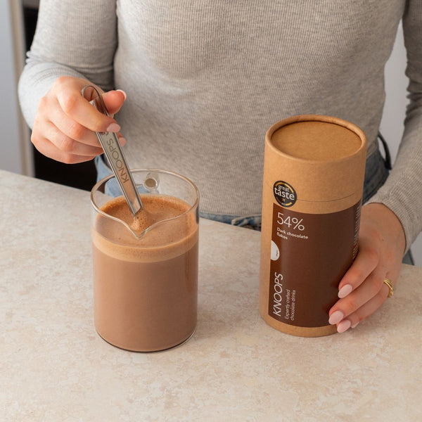 Person holding a glass of chocolate milkshake with a spoon, next to a cardboard container labeled 'Koko's' on a light surface.